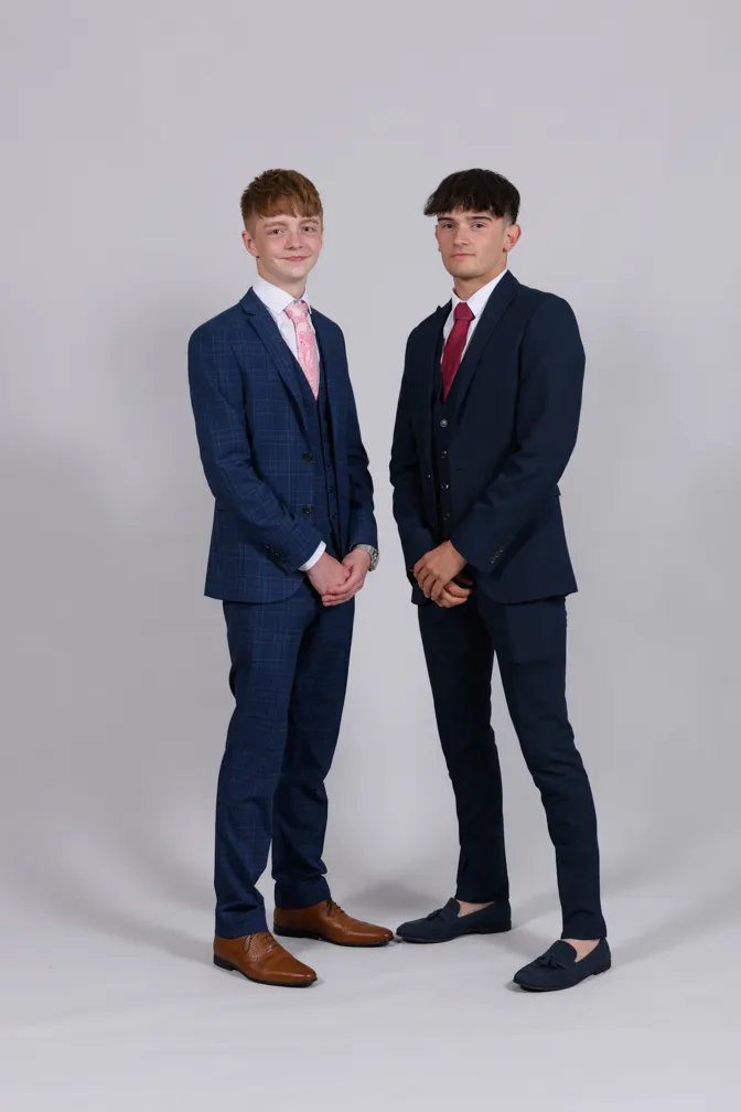 Two young men standing side by side wearing dark suits and dress shoes against a plain light gray background.