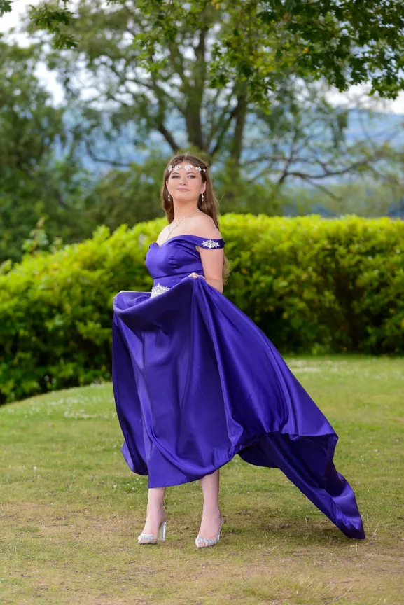 Young woman outdoors wearing an off-shoulder purple satin gown holding the skirt with one hand, surrounded by greenery.