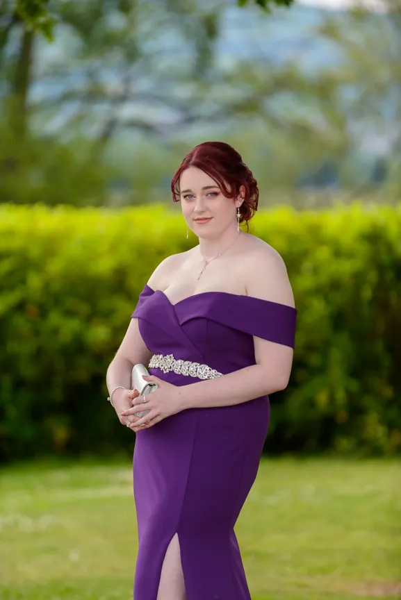Young woman with red hair in an off-shoulder purple dress holding a silver clutch, standing outdoors with blurred green foliage background.