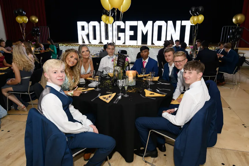 Group of young people dressed in formal attire sitting around a decorated table at an event with illuminated 'ROUGEMONT' letters and black and gold balloons in the background.