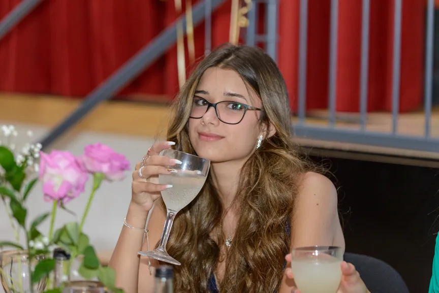 Young woman with long curly hair and glasses holding a glass of white wine or champagne at a social event with pink flowers in the background.