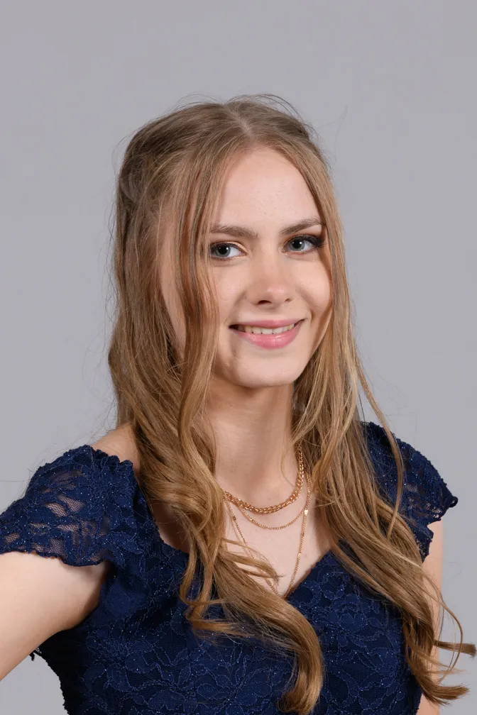 Smiling young woman with long wavy blonde hair wearing a navy blue lace dress and gold layered necklaces.