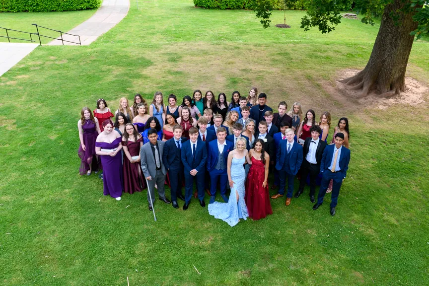 Group of young adults dressed in formal attire standing together on green grass under a large tree.