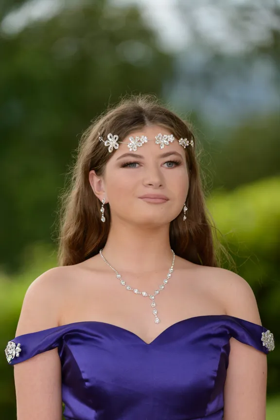 Young woman wearing an off-the-shoulder purple dress and floral silver jewelry against a blurred greenery background.