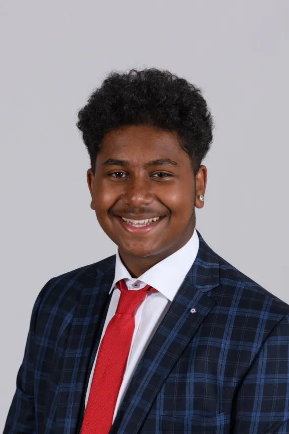 Smiling young man wearing a blue plaid suit jacket, white shirt, and a red tie against a plain gray background.