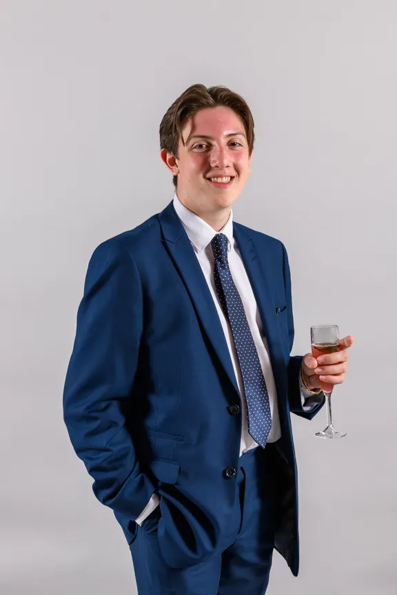 Young man in a blue suit and polka dot tie smiling and holding a glass of wine.