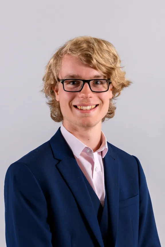 Smiling young man with glasses and blond curly hair wearing a navy blue suit and light pink shirt against a gray background.