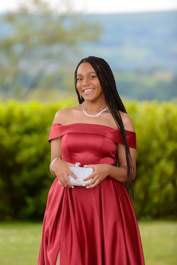 Young woman with long braided hair wearing an off-shoulder red satin gown holding a silver clutch outdoors.