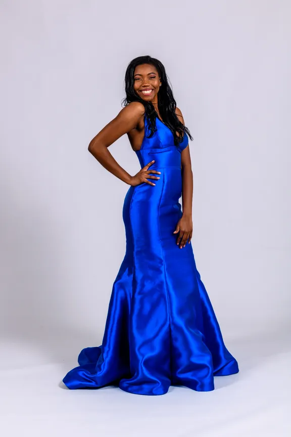 Woman in a vibrant blue satin mermaid-style gown smiling with one hand on her hip against a plain light background.
