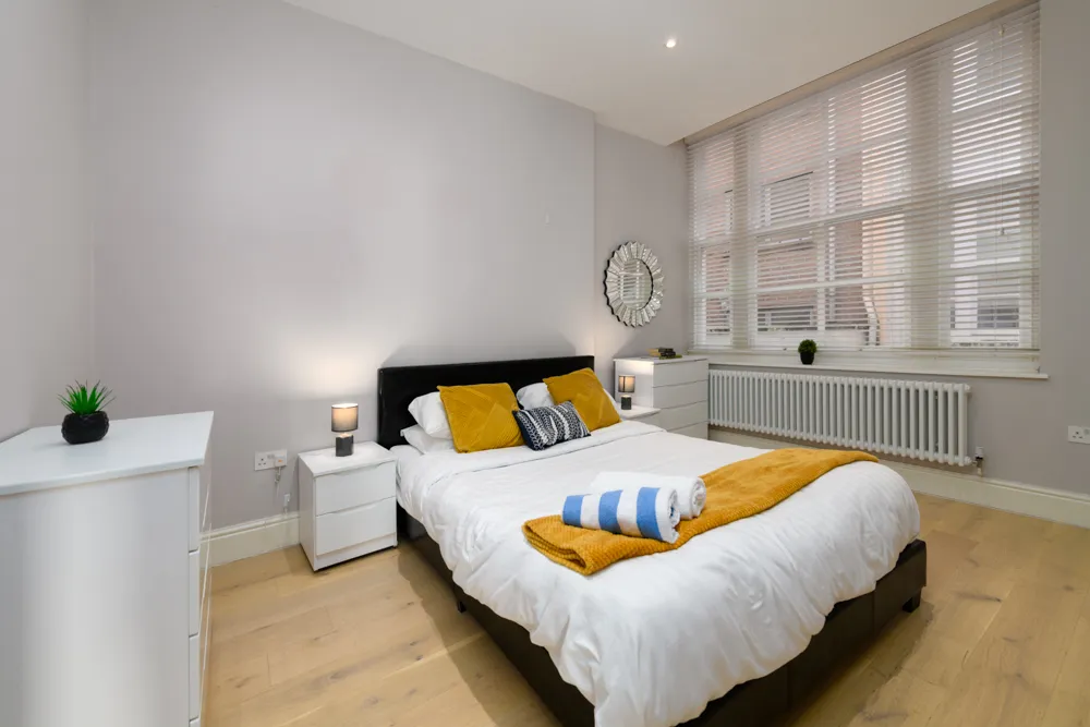 Modern bedroom with a black bed frame, white bedding, mustard-yellow pillows and throw, white furniture, large window with blinds, and wooden floor.