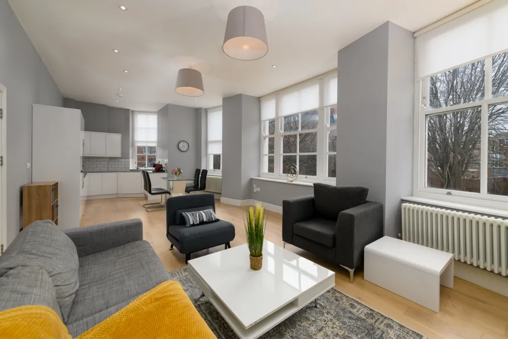 Modern living room with gray sofa, black armchairs, white coffee table, and large windows with white blinds overlooking trees.