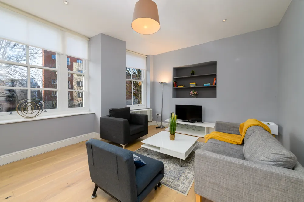 Modern living room with gray walls, large windows, a gray couch with a yellow throw, two dark armchairs, a white coffee table with a plant, and a TV on a low white stand under built-in shelves.