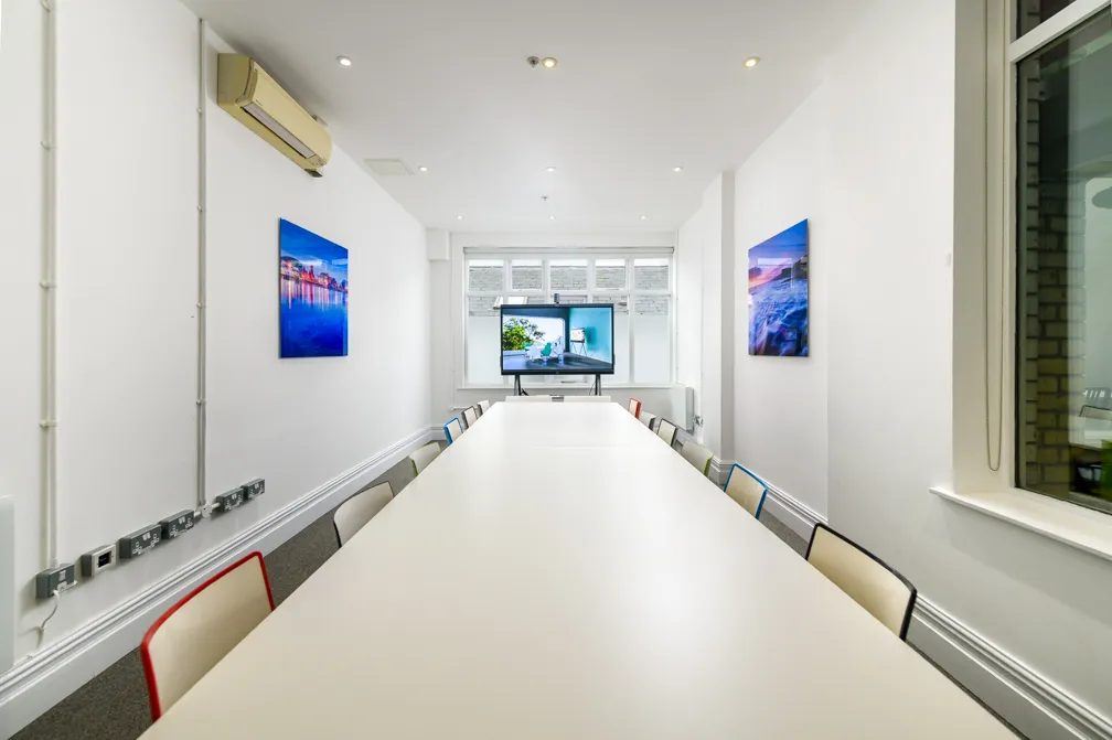 Long white conference table surrounded by chairs with multicolored backs in a bright modern meeting room with a TV screen and wall art.