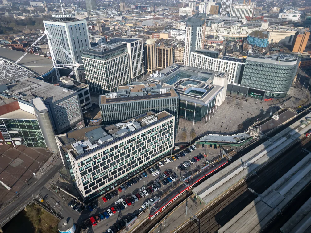 Aerial view of a cityscape showing modern office buildings, a large public square, parked cars, and a red and white train at a railway station.