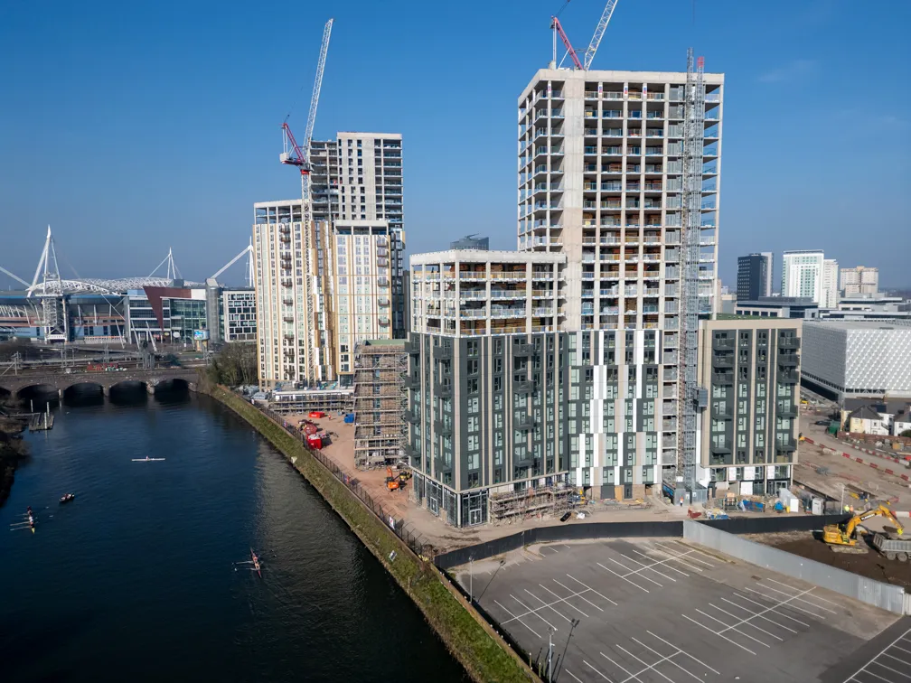 Aerial view of modern high-rise residential buildings under construction near a river with cranes and a parking lot under clear blue sky.