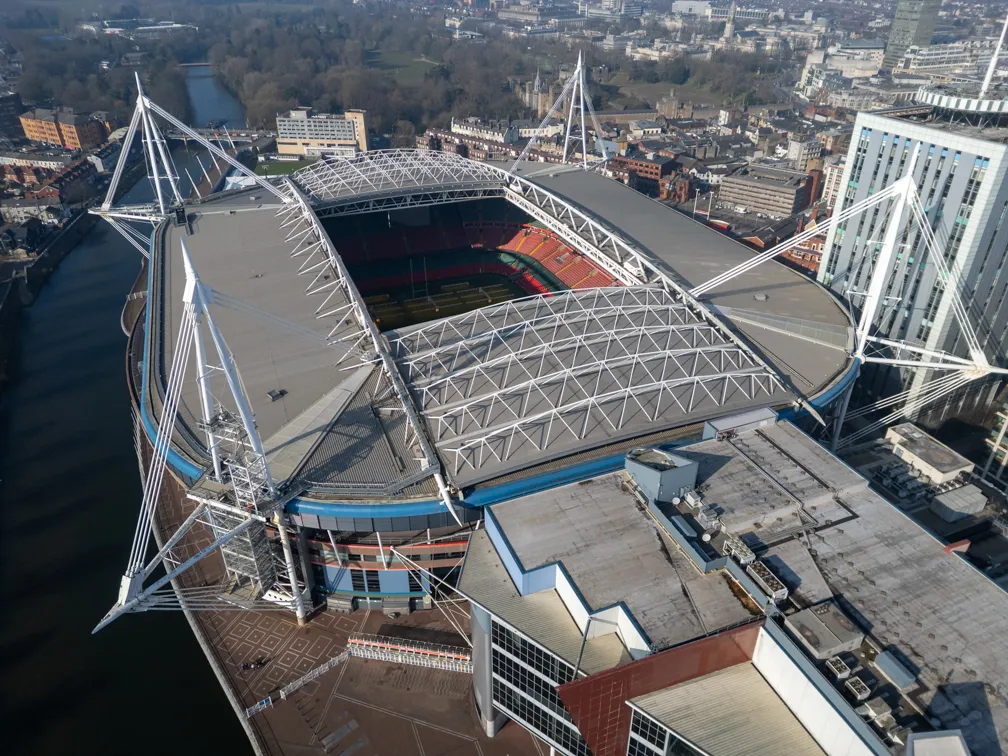 Aerial view of a large stadium with a partially open retractable roof beside a river and city buildings.