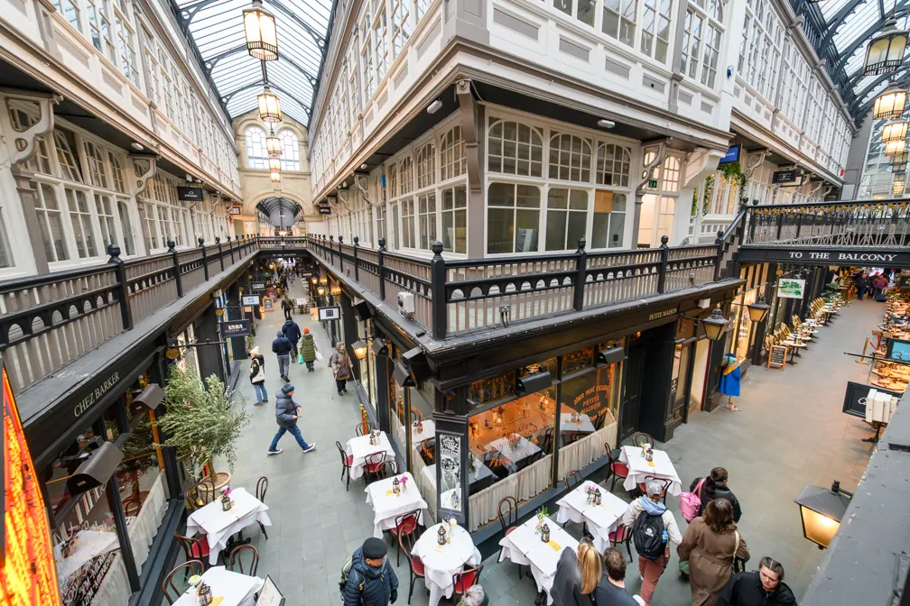 Indoor shopping arcade with a glass roof, featuring small cafes with white tablecloths and people walking along the central corridor.