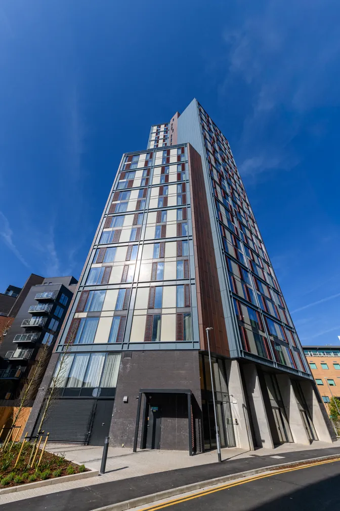 Modern tall residential building with glass and metal panels under a clear blue sky, viewed from street level.
