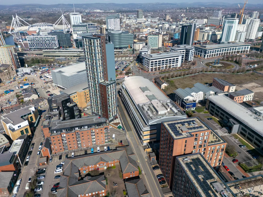 Aerial view of a cityscape featuring modern high-rise buildings, parking lots, roads, and distant hills under a clear sky.