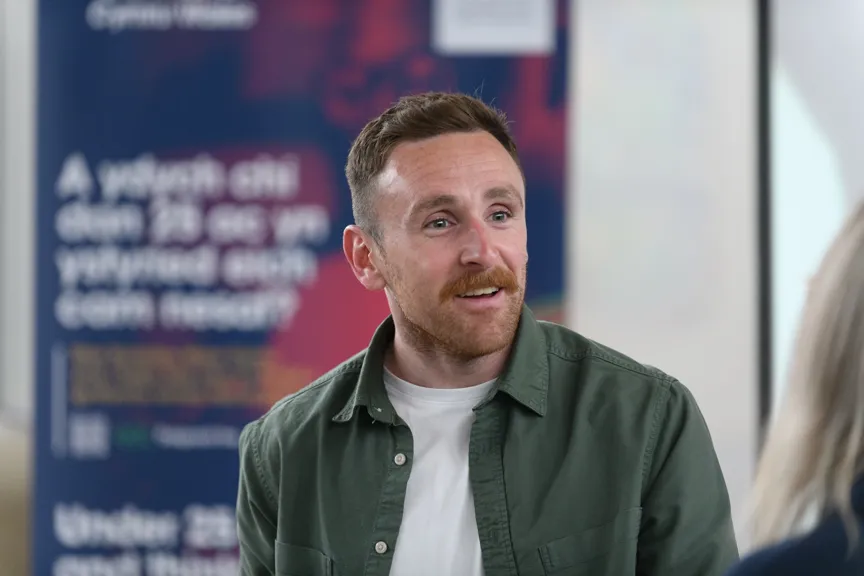 Smiling man with short brown hair and mustache wearing a green jacket and white shirt, talking to a person with blonde hair.