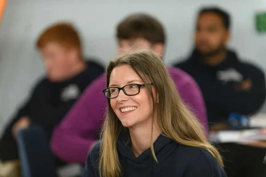 Smiling woman with glasses and long hair in a casual setting with people in the background.