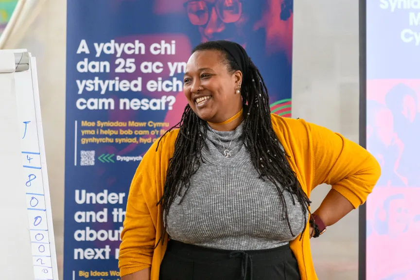 Smiling woman with long dreadlocks in a gray shirt and mustard cardigan standing in front of posters and a whiteboard during a presentation.