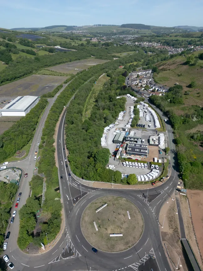 Aerial view of a large roundabout with roads and a vehicle storage area filled with white vehicles surrounded by greenery and hills in the background.