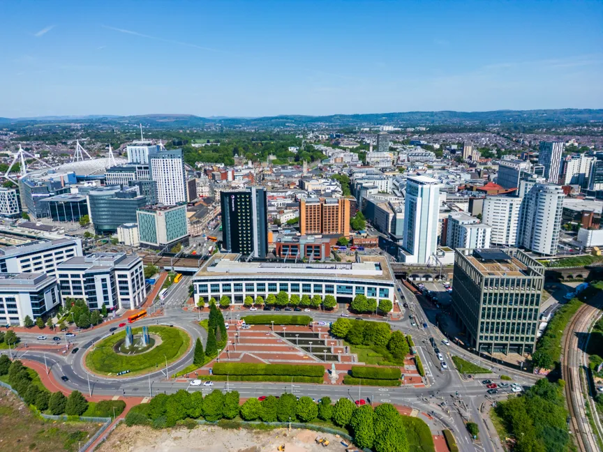 Aerial view of a cityscape with modern buildings, a roundabout with a sculpture in the center, and green trees under a clear blue sky.