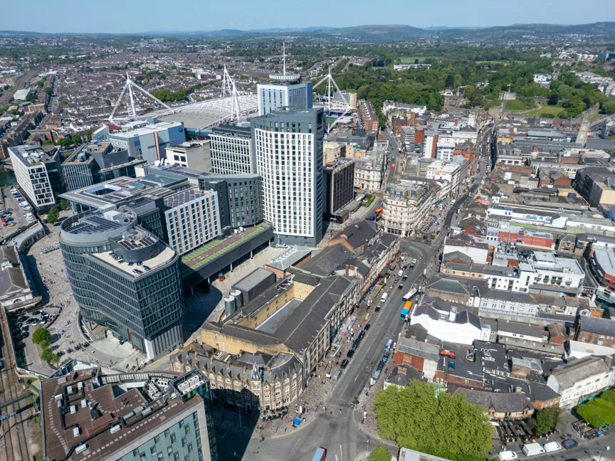 Aerial view of an urban cityscape featuring modern buildings, a stadium with white roof supports, and surrounding roads filled with vehicles.