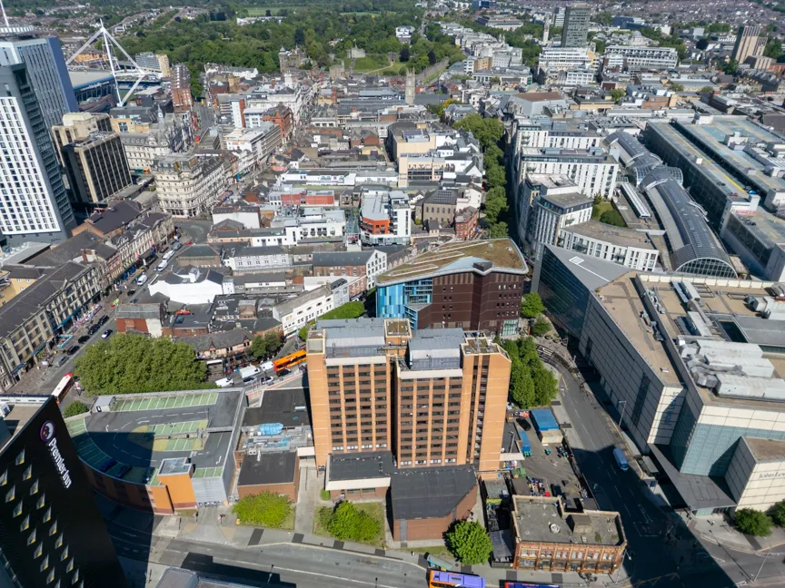 Aerial view of an urban cityscape with a mix of tall and mid-rise buildings, roads, trees, and buses under a clear sky.