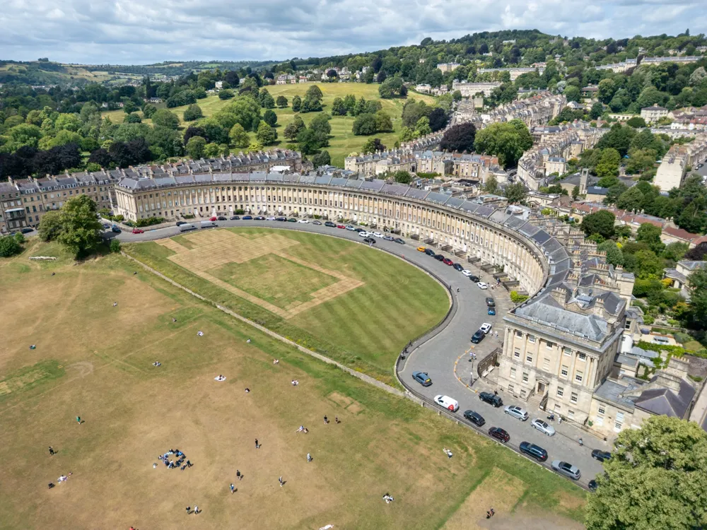 Aerial view of the Royal Crescent, a row of classical Georgian townhouses curving around a large open green lawn, with parked cars and people scattered on the grass in Bath, England.