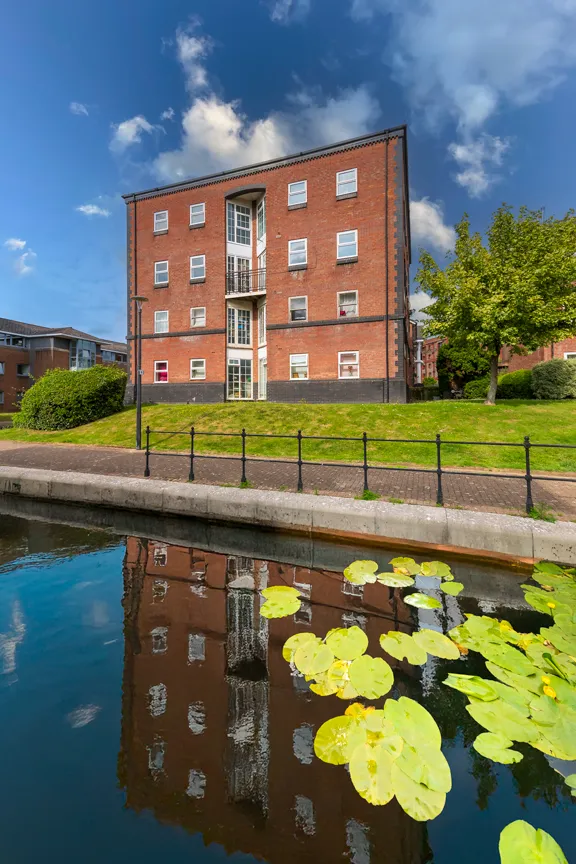 Four-story red brick apartment building reflected in a canal with yellow lily pads in the foreground under a blue sky with clouds.