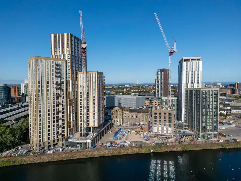 Aerial view of a modern urban waterfront area under construction with tall apartment buildings, cranes, and a canal in the foreground.