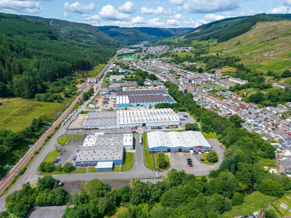 Aerial view of an industrial area with warehouses and parking surrounded by a residential neighborhood and green hills under a partly cloudy sky.