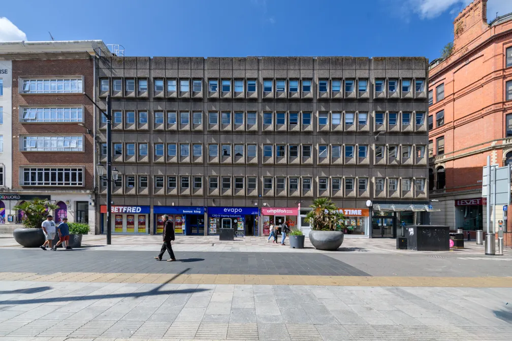 A concrete multi-story commercial building with small rectangular windows above shops including Betfred, Evapo, Amplifon, and Time, under a blue sky.