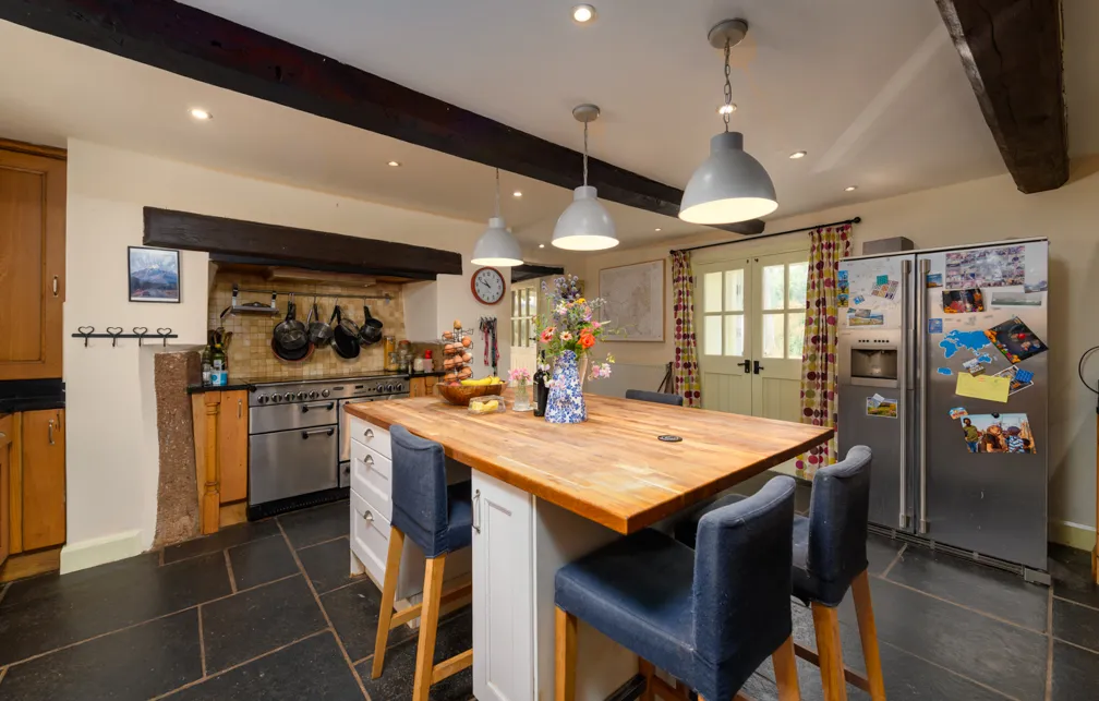 Cozy kitchen with wooden island countertop, blue cushioned stools, hanging pots above stainless steel stove, and stainless steel fridge with magnets.