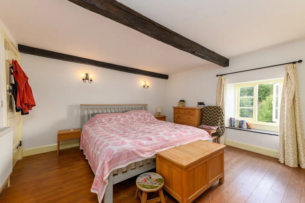 Cozy bedroom with white walls, exposed wooden beams, a bed with pink floral bedding, wooden furniture, and an open window with curtains letting in natural light.