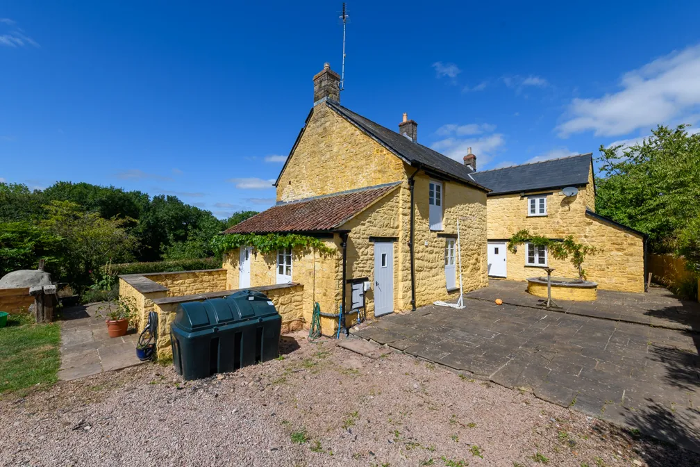 Yellow stone house with white doors and windows under a clear blue sky, surrounded by greenery and a paved patio.