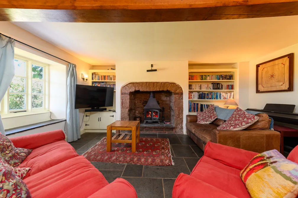 Cozy living room with red sofas, a wood-burning stove in a stone fireplace, bookshelves, a wooden coffee table, and a large window with light blue curtains.