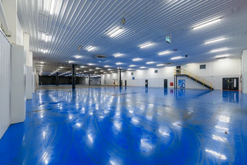 Spacious industrial warehouse interior with shiny blue epoxy floor, white walls, ceiling lights, and a yellow staircase.