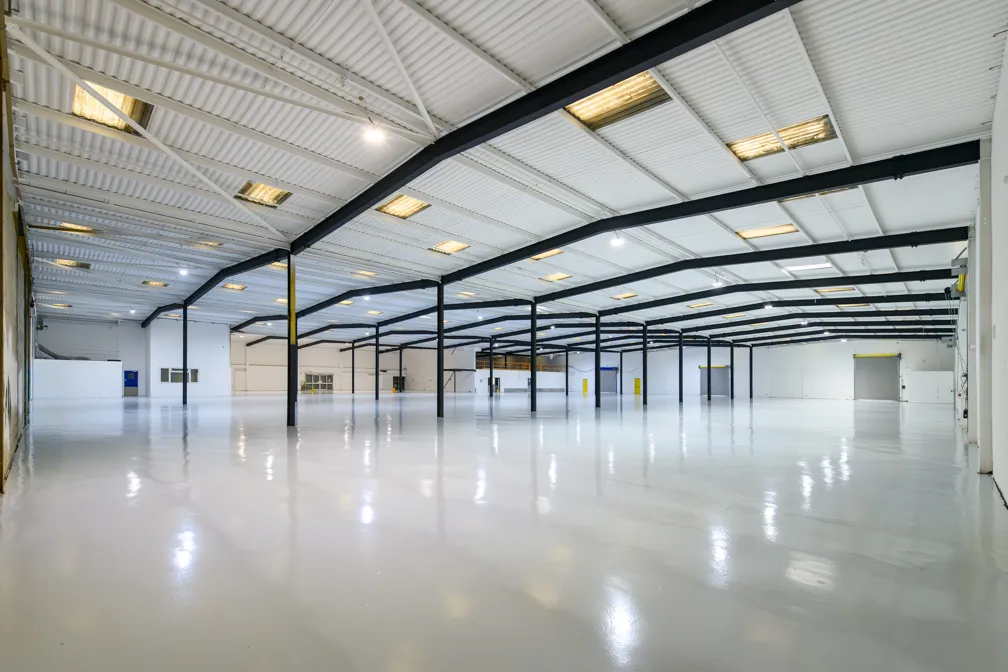 Large empty industrial warehouse with polished white floor and black steel beams supporting a white corrugated metal ceiling.
