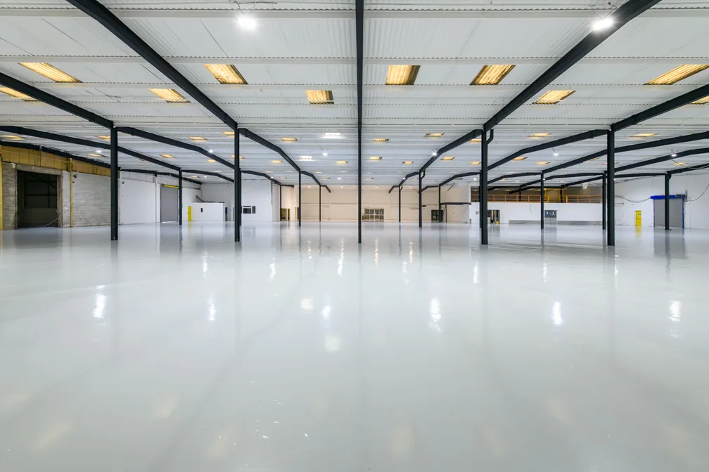 Wide view of a large empty industrial warehouse with glossy white floor, black support beams, and bright ceiling lights.