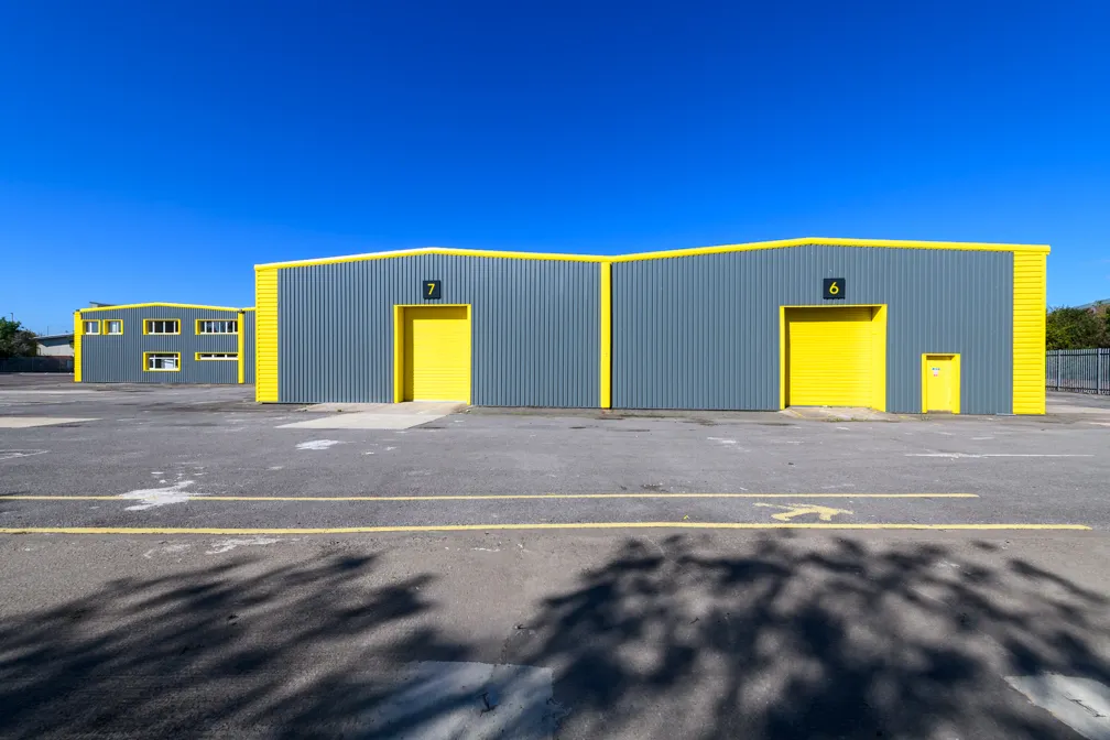 Two adjacent grey warehouse buildings with bright yellow trim, doors numbered 6 and 7, under a clear blue sky.