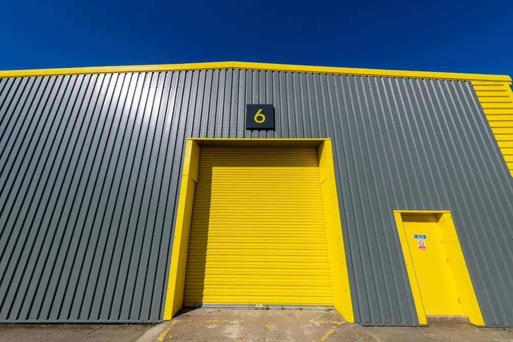 Industrial building with gray metal siding and a large yellow roll-up door numbered 6 next to a smaller yellow door under a clear blue sky.
