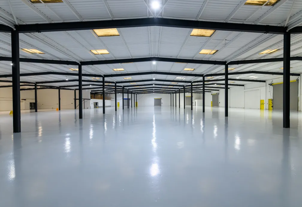 Spacious empty industrial warehouse with white polished floor, black ceiling beams, and bright overhead lights.