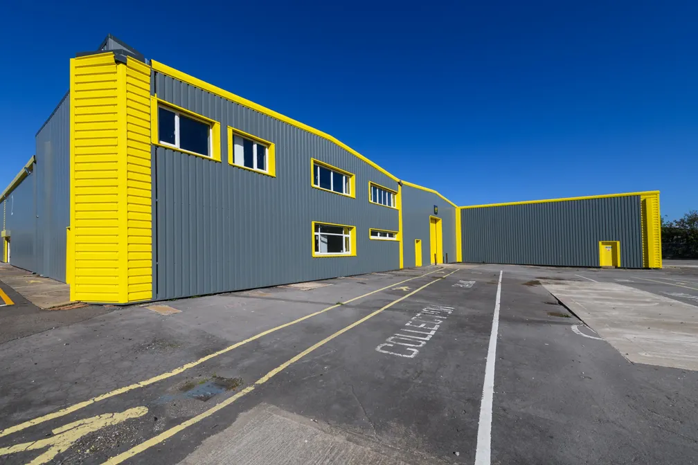 Large industrial warehouse with gray walls and yellow trim under a clear blue sky.
