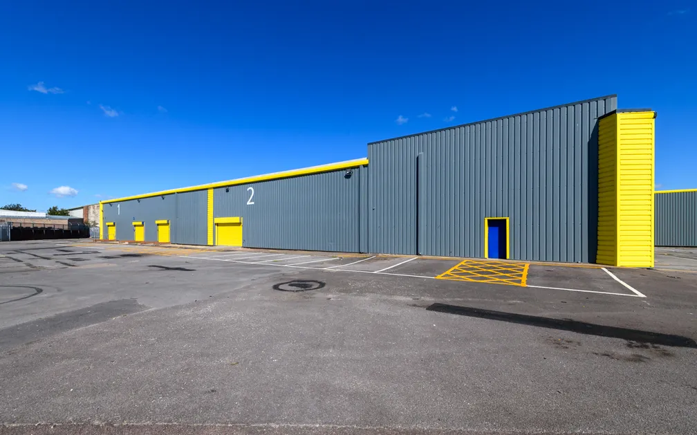 Gray industrial warehouse buildings with yellow accents and numbered doors under a blue sky.