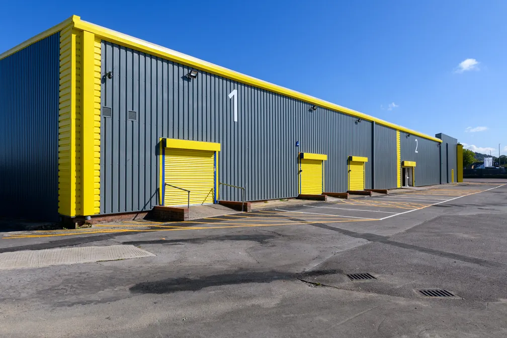 Large industrial warehouse with grey walls and bright yellow doors and trim, marked with white numbers 1 and 2 under a clear blue sky.