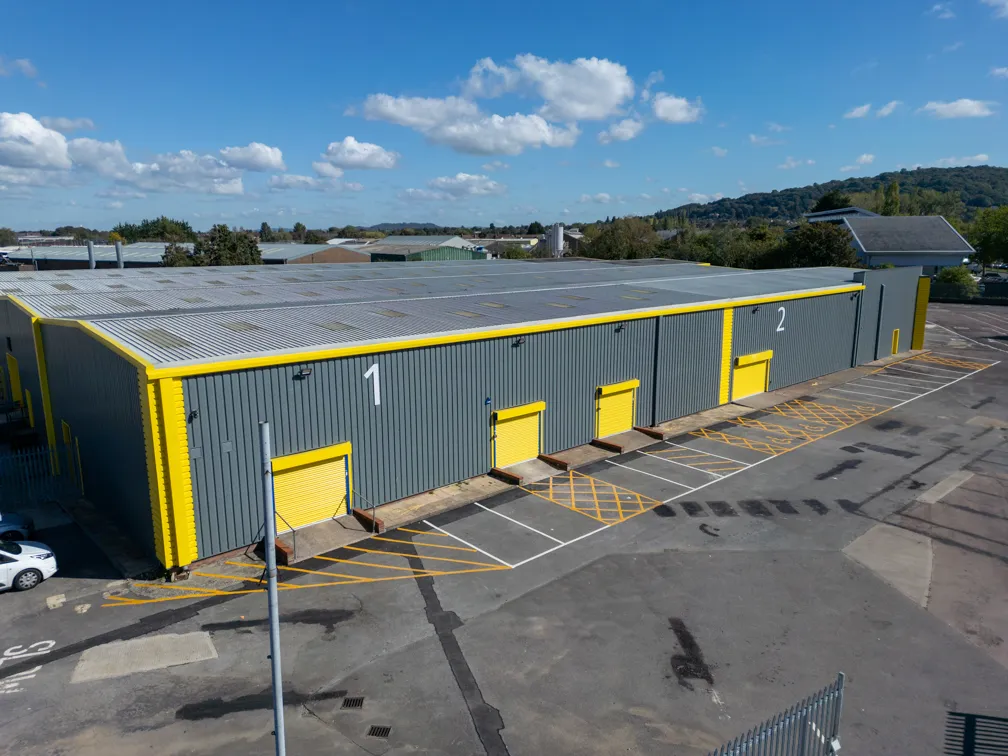 Aerial view of two large industrial warehouse units with gray walls and yellow accents, featuring numbered loading doors 1 and 2 under a partly cloudy sky.