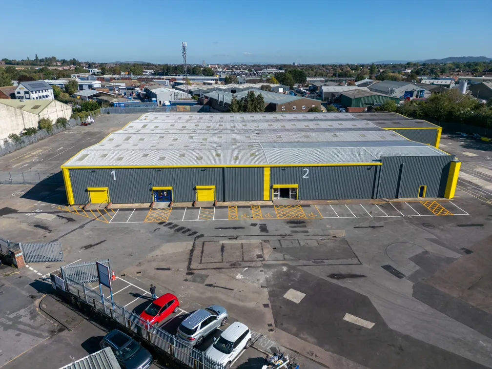 Aerial view of a large industrial warehouse with numbered sections 1 and 2, surrounded by a parking area and several parked cars.
